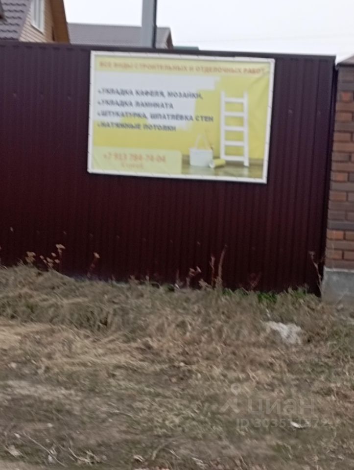 На фотографии виден забор из металлических панелей темного цвета, который обрамляет участок. На заборе размещен рекламный баннер с ярким фоном, на котором указаны услуги по укладке кафеля, мозаики, а также отделке стен и потолков. Цвета баннера — желтый и белый, текст черного цвета. Структура самого забора выполнена из металлических листов, что придает ему современный вид. Внизу видны сухие травы и незначительные растительности, что создает ощущение запущенности территории.