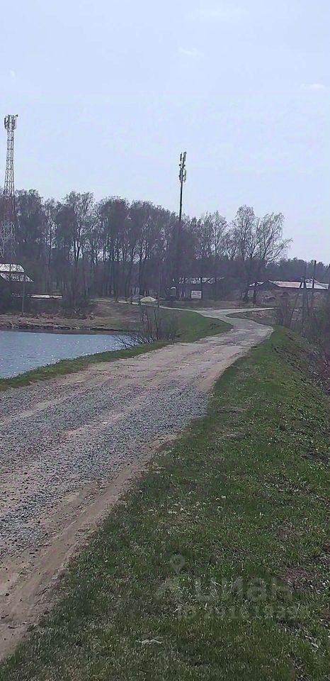 На фотографии изображена грунтовая дорога, которая извивается вдоль водоема. По обеим сторонам дороги видны участки зелёной травы и кустарников. Вдали можно заметить несколько деревьев, создающих тень и придающих ландшафту природный вид. Дорога имеет каменистую поверхность и, похоже, используется для проезда. На заднем плане видны строения, однако их детали неразличимы. Общее впечатление от изображения создает спокойная и уединенная атмосфера, характерная для загородной местности.