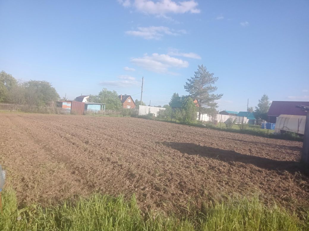 На изображении представлено пустое поле с обработанной землёй, готовой для посадки. Почва имеет тёмно-коричневый цвет и выглядит рыхлой, что свидетельствует о свежей обработке. Вдали видны несколько строений, включая дома с крышей разных оттенков, а также хозяйственные постройки. На заднем плане располагаются деревья и кустарники, создающие естественный фон. Участок окружён деревянным забором, что придаёт ему уединённость. Общая атмосфера пространства спокойная и сельская, идеально подходящая для ведения дачного хозяйства.