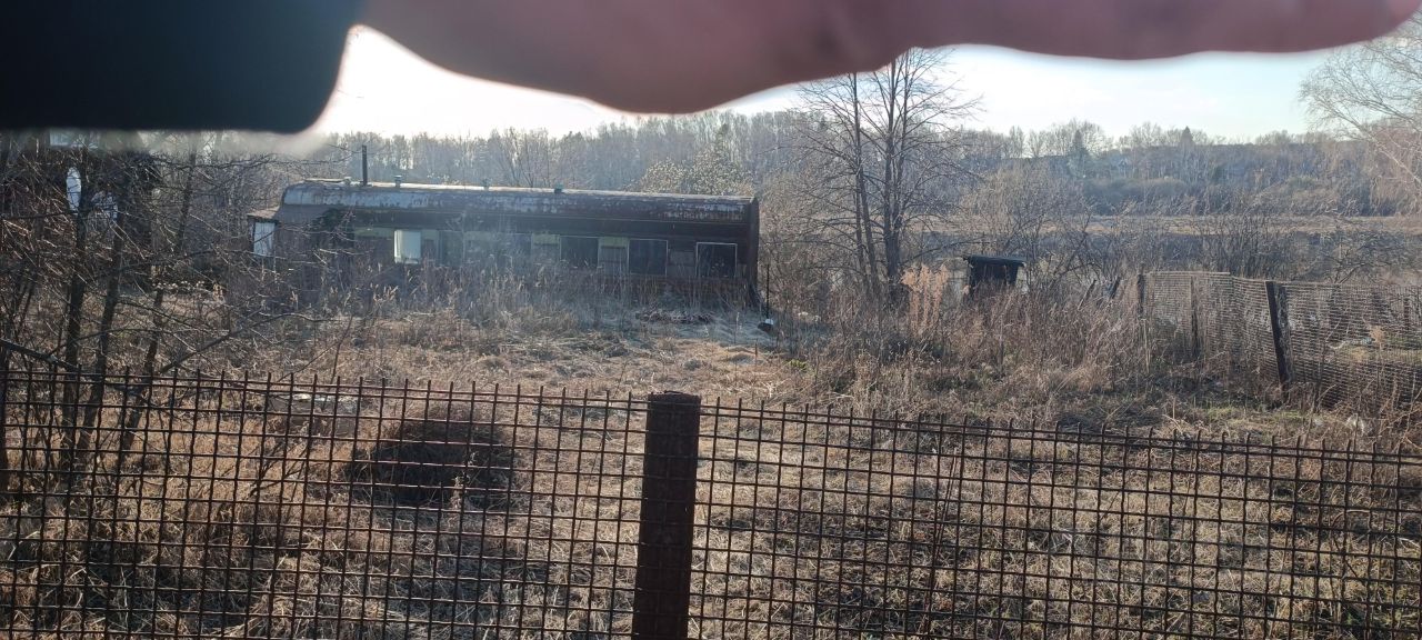 На фотографии виден заброшенный вагон, окруженный высокой травой и кустарниками. Стены вагона имеют ржавчину и следы старения, окна заколочены. Перед вагоном расположена металлическая сетка, которая ограничивает доступ к территории. Вдали видны деревья, что создает ощущение уединенности. Общая цветовая палитра включает землистые и ржавые оттенки, что подчеркивает запустение места. Пространство кажется заброшенным, с отсутствием человеческого присутствия и ухоженности.