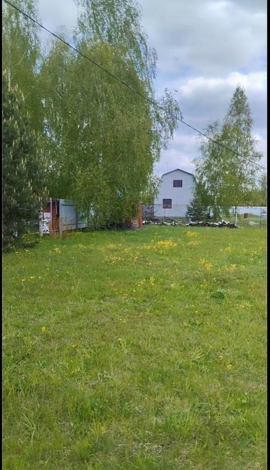 На фотографии виден просторный участок земли с зелёной травой и желтыми цветами. В центре композиции расположены высокие деревья, создающие тенистую атмосферу. На заднем плане виднеется строение с простым архитектурным дизайном, выполненное из светлого материала. Окна здания имеют прямоугольную форму, а крыша двускатная. Участок выглядит ухоженным, с минимальными постройками, что подчеркивает его природную красоту и возможность для дальнейшего использования. Простор открывает множество вариантов для ландшафтного дизайна и создания уютного пространства для отдыха.
