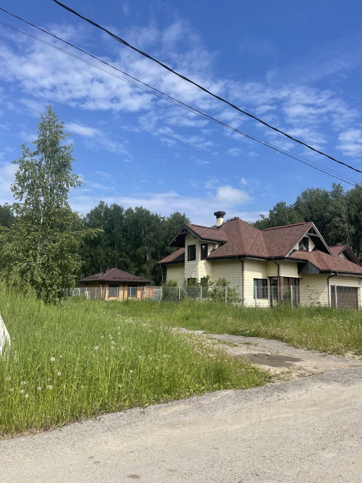 Здание выполнено в загородном архитектурном стиле с элементами классической архитектуры. Крыша с черепицей имеет сложную форму, а фасад отделан светлыми деревянными панелями. Видны большие окна с декоративными наличниками, создающими ощущение уюта. Справа расположена отдельная постройка, возможно, гараж или хозблок. Открытая территория вокруг дома обрамлена зелеными насаждениями и высоким травяным покровом, что подчеркивает природное окружение. Цветовая гамма здания включает светлые и коричневые оттенки, гармонирующие с окружающим ландшафтом.