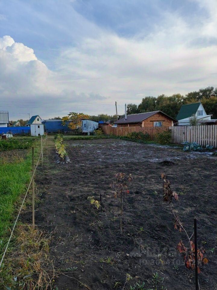 На фото представлено пустое земельное пространство с подготовленной почвой, готовой для посадки. Вдали видны несколько садовых участков с небольшими домиками, выполненными из дерева и окрашенными в разные цвета. По периметру участка установлены столбы, возможно, для ограждения. На земле видны небольшие растения, что указывает на начальную стадию озеленения. Общая атмосфера участка создает ощущение уединения и готовности к дальнейшему развитию, с акцентом на сельскую жизнь и садоводство.