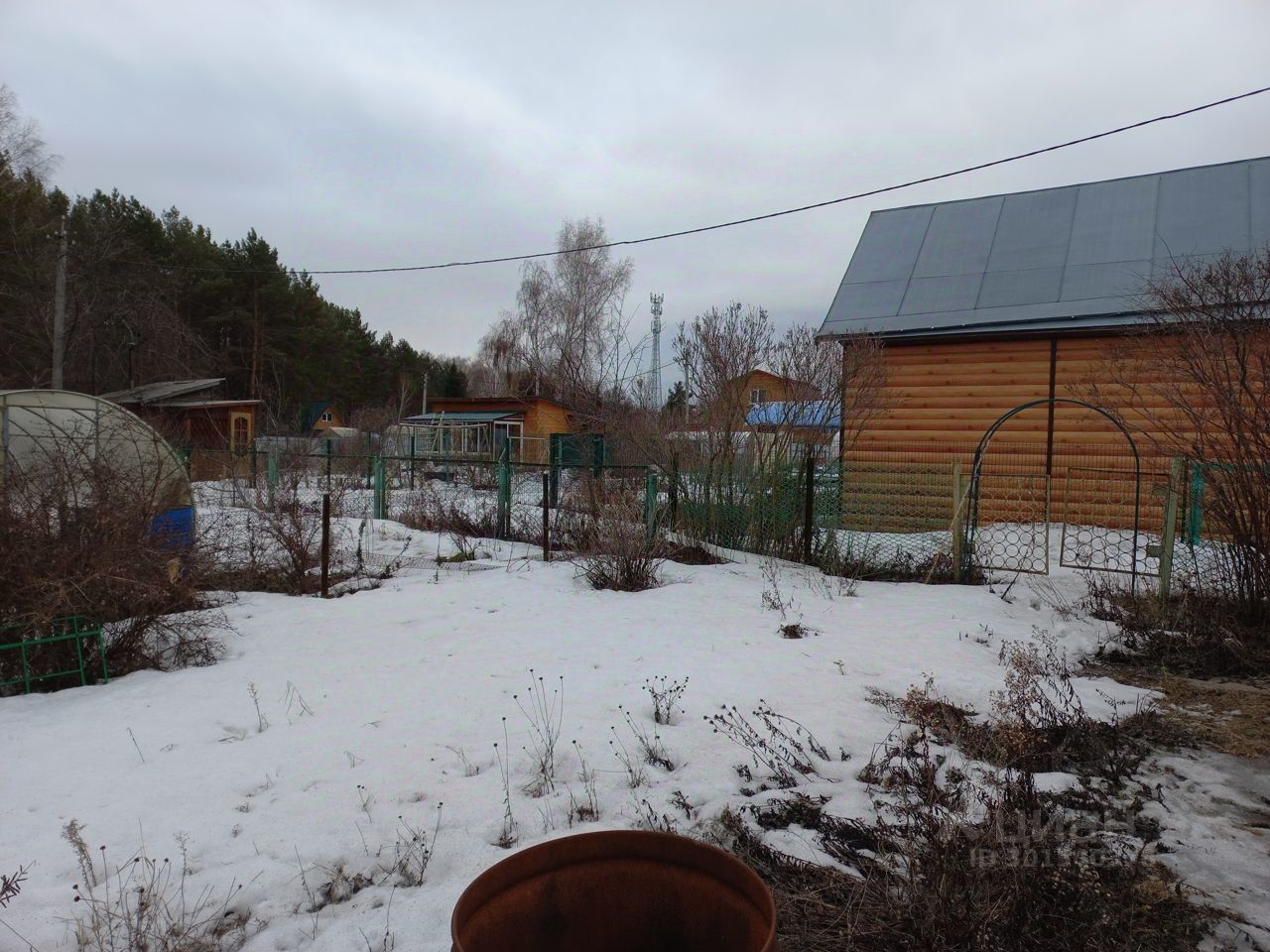 На фотографии виден загородный участок с несколькими постройками. Основные элементы архитектуры представляют собой деревянные дома с крышей, покрытой металлическими листами. Структурные особенности включают заборы из металлической сетки и арку, ведущую в сад. Участок частично покрыт снегом, вокруг видны кустарники и сухая трава. Основные цвета — коричневый от древесины, серый от крыши и зеленый от забора. Завершает картину неухоженная территория с элементами ландшафтного дизайна, создающая атмосферу уединения и спокойствия.