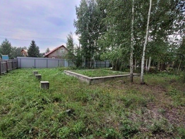На фотографии виден участок земли с незавершённым строительством. Центральное место занимает бетонный фундамент прямоугольной формы, окружённый зелёной травой и кустарниками. По периметру участка расположены деревянные столбы, возможно, для установки забора. На заднем плане видны деревья, создающие тень, а также частично виден дом с красной крышей. Общая атмосфера спокойная и уединённая, что делает пространство подходящим для дальнейшего строительства или отдыха на природе.