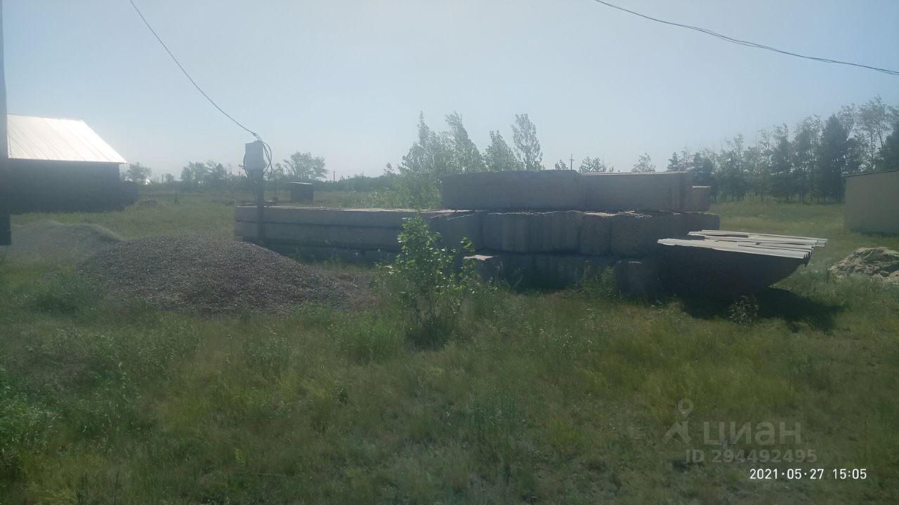 На фотографии видно участок земли с несколькими большими бетонными плитами, аккуратно уложенными друг на друга. Рядом находятся груды гравия и небольшие кустарники, что придаёт пространству естественный вид. Вдалеке виден деревянный строение с металлической крышей. Цвета в основном нейтральные, с преобладанием серых и коричневых тонов. Структура выглядит прочной и долговечной, что может указывать на строительные материалы высокого качества. Пространство открытое и не перегружено объектами, создавая ощущение простора.