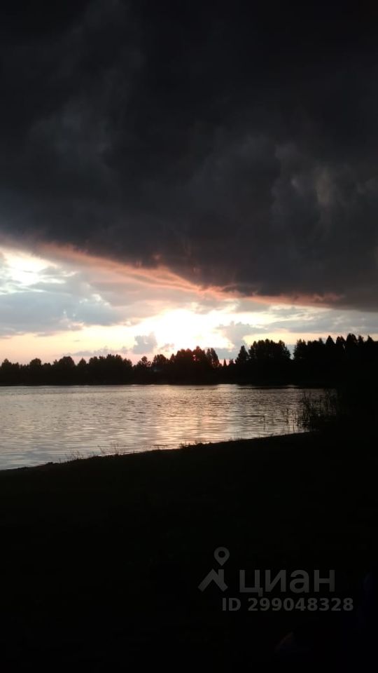 Вид на водоем с отражением заката и облаков, создающих драматичную атмосферу. Гладкая поверхность воды отражает яркие оттенки оранжевого и розового, контрастируя с темными облаками. На заднем плане виднеются силуэты деревьев, подчеркивающие спокойствие и уединение этого места. Вода и небо гармонично сочетаются, создавая живописный пейзаж. Общая цветовая палитра включает глубокие серые, синие и теплые тона, что придаёт сцене особую атмосферу. Это пространство идеально подходит для отдыха и наслаждения природой.