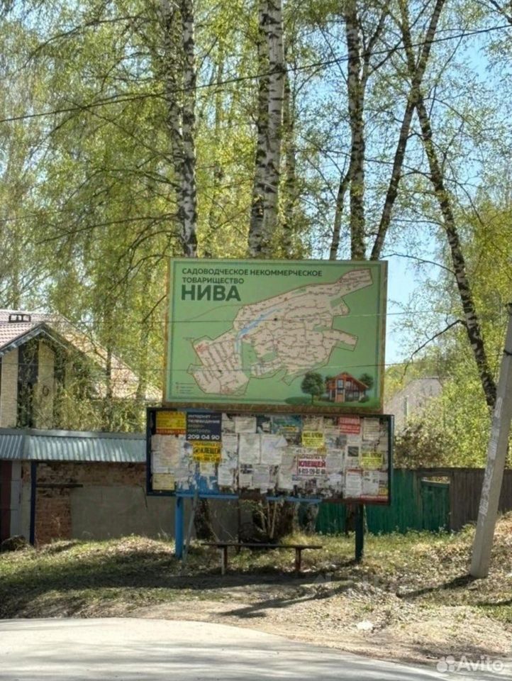 На фотографии представлена информационная доска с картой садоводческого товарищества. Доска выполнена из металла и окрашена в светло-зеленый цвет. На ней изображена схема участков с обозначениями, а также информация о правилах и событиях. В нижней части доски размещены объявления, прикрепленные на бумаге, с текстом на русском языке. Фон составляет зеленая растительность, в частности березы, что создает природный контекст. Структура в левом углу выглядит как одноэтажное здание с крышей из металлочерепицы, выполненное в светлых тонах.