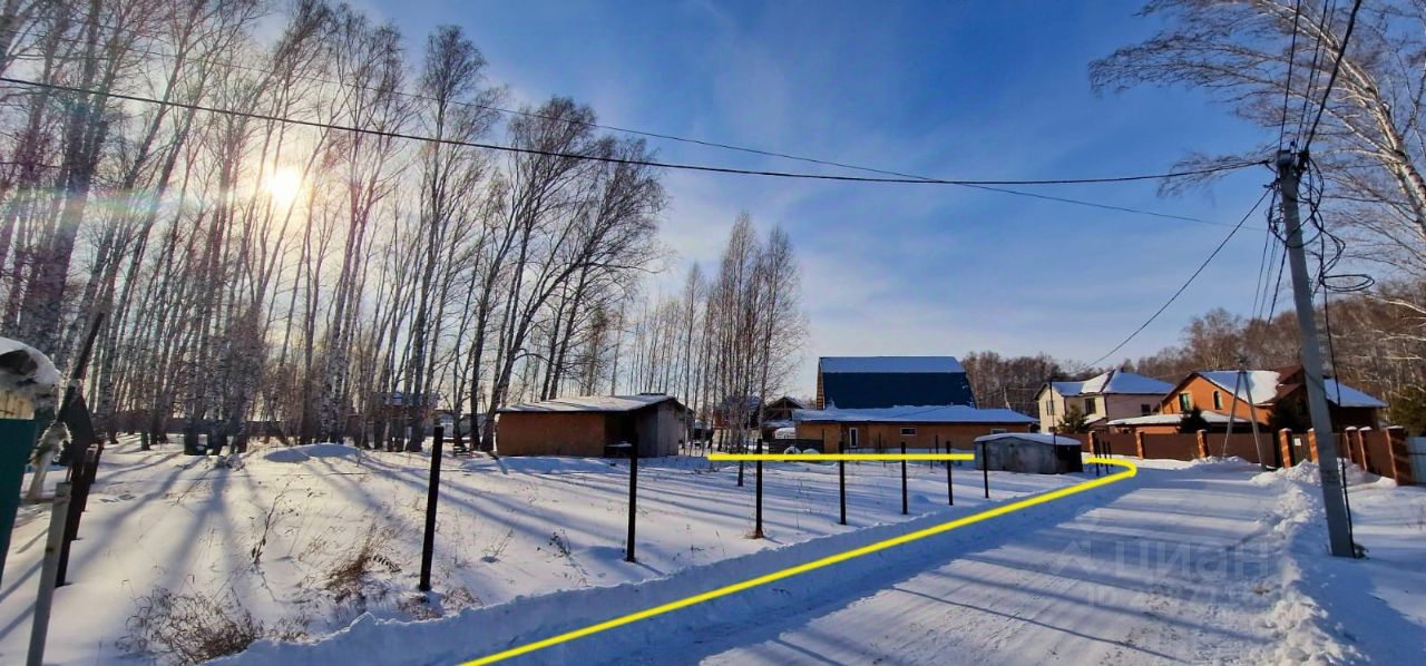 На фотографии видно зимний пейзаж с заснеженной дорогой, окруженной деревьями. Строения представляют собой дома с простыми архитектурными формами, выполненные из кирпича и дерева. Некоторые здания имеют двускатные крыши, что придаёт им традиционный вид. Цвета фасадов варьируются от светло-коричневого до серого, что создает гармоничное сочетание с зимним окружением. Заборы из металлических прутьев разделяют участки, а снежный покров на земле и крыше придаёт сцене спокойствие и уединение. Светлая атмосфера создаётся солнечными лучами, пробивающимися сквозь деревья.