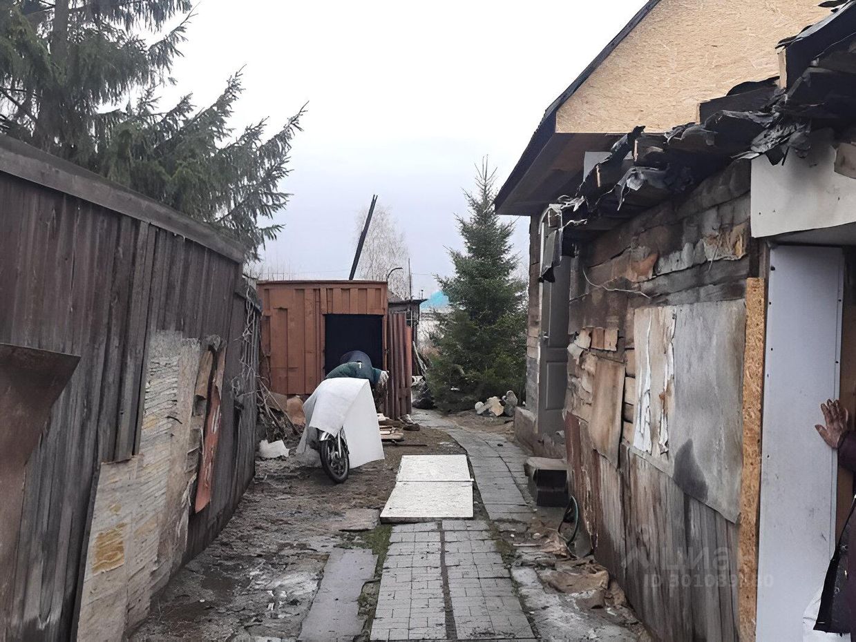 Узкая дорожка, выложенная плиткой, проходит между деревянными заборами. С одной стороны видна стена дома с обшивкой из камня и поврежденной крышей. На стене имеются следы старой отделки и различные материалы, использованные для ремонта. С другой стороны расположена деревянная постройка, частично закрытая от взглядов. На дорожке стоит мотоцикл, накрытый белым материалом. Вдали видны хвойные деревья, добавляющие зелени в эту местность. Общая атмосфера пространства создает ощущение заброшенности и необходимости в ремонте.