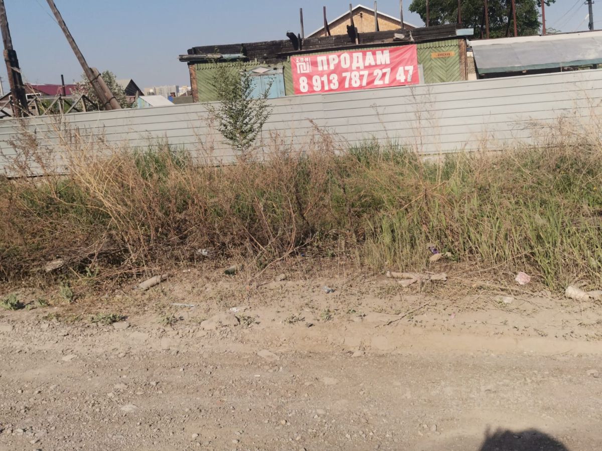 На фотографии виден участок земли, окружённый высоким забором. За забором расположены строения с различными архитектурными элементами, включая деревянные и металлические конструкции. Виден знак с надписью "Продаю", что указывает на наличие предложения о продаже. Растительность вокруг участка неухоженная, с высокой травой и кустарниками. Поверхность земли состоит из грунта и мелких камней, что создаёт неформальную атмосферу. Общий вид участка демонстрирует потенциальные возможности для застройки или использования в коммерческих целях.