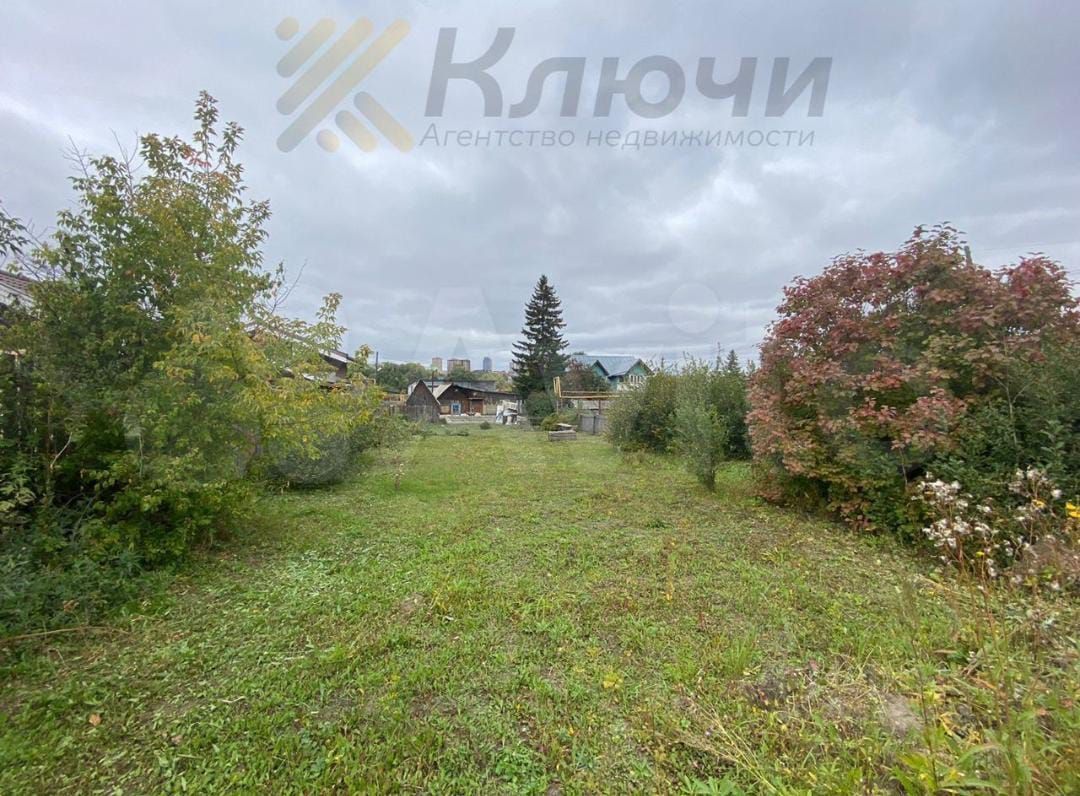 На фотографии виден просторный участок земли с зелеными травами и кустарниками. Участок окружен деревьями, создающими уютную атмосферу. Вдалеке можно заметить несколько строений, которые имеют традиционный дачный стиль. Разнообразные растения добавляют цвет и текстуру, а также создают естественные границы между участками. Общая картина передает ощущение спокойствия и уединения, подходящего для загородной жизни. Цвета в основном натуральные, с преобладанием зеленого и коричневого, что подчеркивает природную красоту этого места.