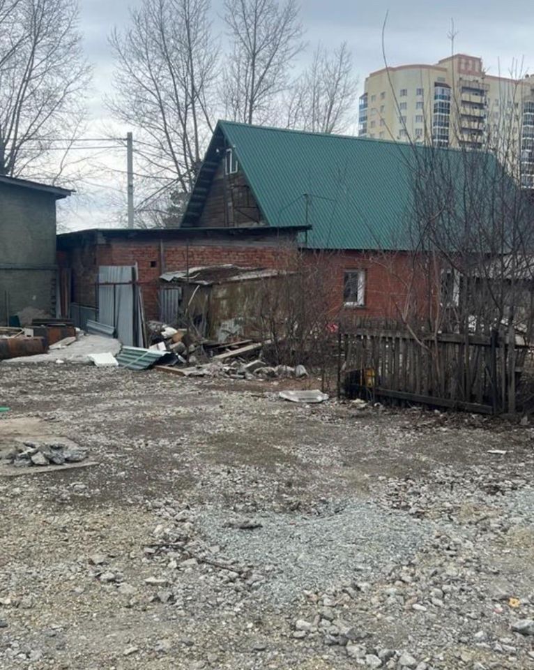 На фото представлено строение с металлической крышей зеленого цвета и кирпичными стенами. Стены частично обветшали, видны следы старой отделки. Внешний вид здания указывает на его возраст и возможную необходимость ремонта. Вокруг строения разбросаны строительные материалы и мусор, что создает ощущение заброшенности. Площадка перед зданием состоит из гравия и земли, с небольшими участками растительности. Ограждение из деревянных досок добавляет rustic-элемент в общий вид. Общая атмосфера пространства выглядит неухоженной и требует внимания.