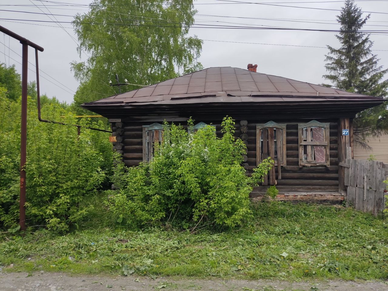 Деревянный дом в традиционном русском стиле с характерной бревенчатой конструкцией. Крыша имеет наклонную форму и покрыта металлическим материалом. Фасад украшен деревянными рамами окон с элементами резьбы, некоторые окна закрыты ставнями. Вокруг здания видны густые зеленые кустарники, придающие уединенность. Цвета фасада естественные, с оттенками коричневого и зеленого. Стены выглядят немного изношенными, что подчеркивает историческую ценность постройки. Общий вид создает атмосферу деревенского уюта и традиционной архитектуры.