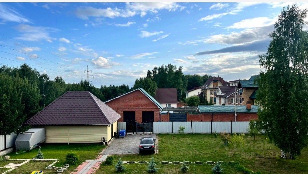 На фотографии представлено пространство с несколькими зданиями, выполненными в разных архитектурных стилях. Основное строение имеет кирпичную отделку и двускатную крышу, что придаёт ему современный вид. Рядом расположено более старинное здание с крышей в форме полусферы. Внутреннее пространство окружено ухоженным газоном и дорожками из плитки. Видно, что во дворе есть парковка для автомобиля. Цветовая палитра включает теплые оттенки кирпича и нейтральные тона стен. Ограда из белых панелей создает ощущение уединения и защищенности.