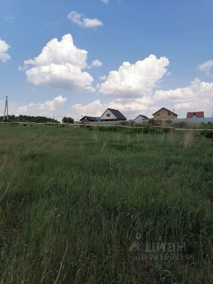 На фотографии виден участок земли с высокой травой и несколькими домами на заднем плане. Архитектурный стиль домов разнообразен: один из них имеет двускатную крышу с черепицей, другой — с плоской крышей и яркими акцентами. Структурные элементы включают деревянные заборы и дорожки, ведущие к домам. Основные цвета — светлые и натуральные, что создает гармоничную атмосферу. Финишные материалы включают дерево и кирпич, придающие постройкам прочность и уют. Пространство открытое и располагает к отдыху на природе.