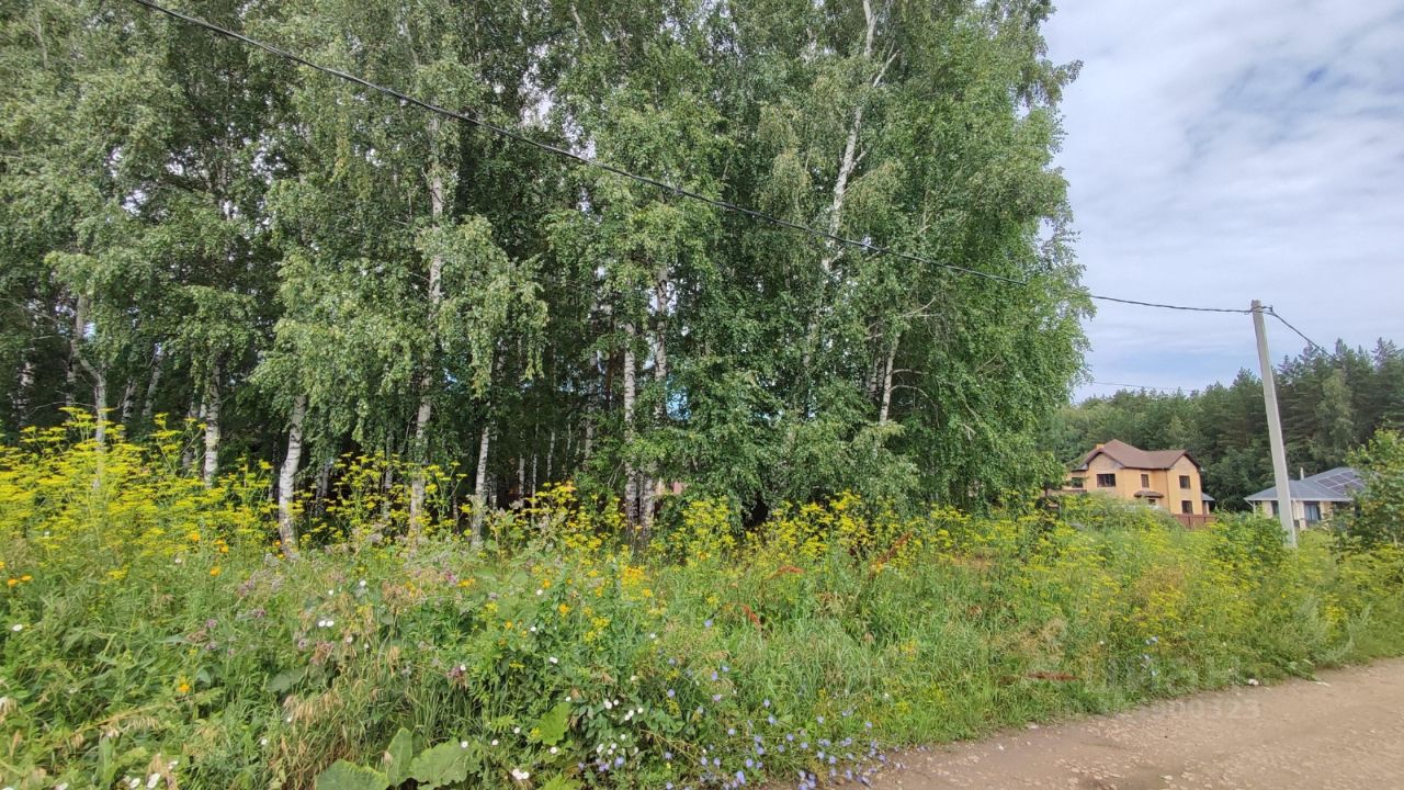 На фотографии виден участок с густыми зелеными деревьями и яркими цветами. Архитектурный стиль построек, расположенных на заднем плане, можно охарактеризовать как современный, с использованием светлых тонов и простых форм. Структурные особенности включают двухэтажные дома с наклонными крышами. Основные характеристики видимой зоны включают ухоженные цветники и травянистые участки, создающие уютную атмосферу. Материалы, использованные в строительстве, выглядят как кирпич и дерево, а цветовая палитра варьируется от светло-коричневого до желтого, гармонируя с природным окружением.