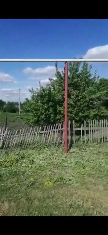 На изображении виден участок земли с металлическим забором, состоящим из вертикальных и горизонтальных перекладин. Забор окрашен в красный цвет и поддерживается металлическими столбами. За ним расположена зелёная растительность, включая кусты и деревья. Площадка выглядит ухоженной, с аккуратно подстриженной травой. На заднем плане видны линии электропередач и открытое пространство, что создаёт ощущение уединения и спокойствия. Цветовая палитра включает зелёные и коричневые оттенки, подчеркивающие природный ландшафт.