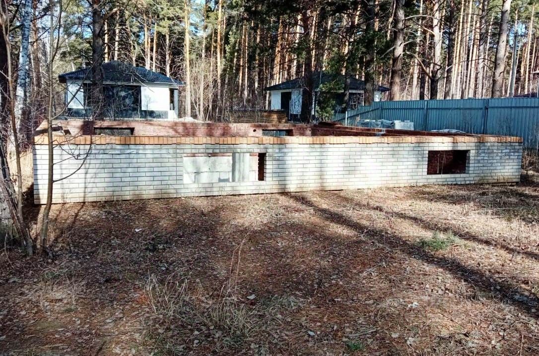 Стена из светлого кирпича с частично открытыми окнами и дверными проемами. На верхней части видны деревянные балки, что указывает на незавершенное строительство. Пространство вокруг выглядит неухоженно, с остатками листвы и травы. Отсутствие крыши и внутренних стен создает ощущение открытости. Стены имеют несколько участков, где кирпич не полностью завершен, что подчеркивает незавершенность проекта. Общий вид демонстрирует прямые линии и простоту архитектурного стиля, характерного для современных загородных домов.