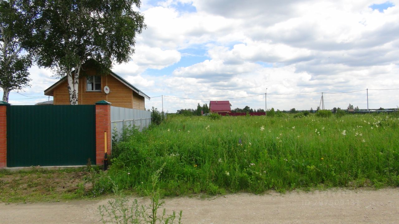 На фотографии представлено загородное строение в деревянном архитектурном стиле с наклонной крышей. Видны элементы, такие как окна с простыми рамами и антенна на крыше. Строение окружено зеленым забором из кирпича и металлическими панелями. Перед домом располагается участок с высокой травой и дикими цветами, создавая естественный ландшафт. Основные цвета — теплые оттенки дерева и зеленый забор, что придает уютный вид. Просторный участок позволяет для дальнейшей планировки и использования, создавая возможность для садоводства или отдыха на свежем воздухе.