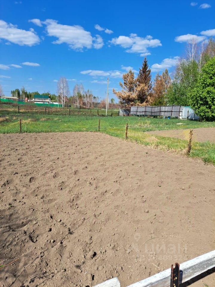 На фотографии представлено открытое пространство с обработанным земельным участком. Земля имеет рыхлую текстуру, готовую для посадки, с ровными рядами. Вдали видны зеленые насаждения и ограждение, создающее ощущение уединения. С одной стороны участка расположены деревья, добавляющие природный элемент, а также видно несколько строений, которые могут служить для хранения или других нужд. Общая цветовая палитра включает коричневые и зеленые оттенки, что подчеркивает сельский характер данной местности.