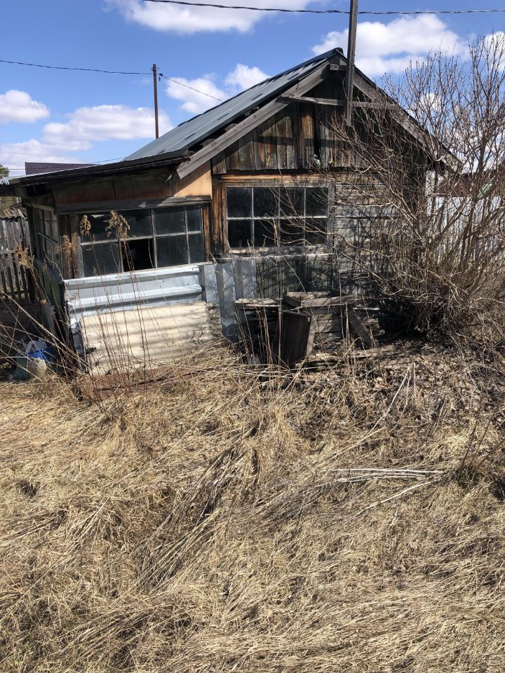 Строение выполнено в простом архитектурном стиле с элементами rustic. Видны деревянные панели и металлические листы, образующие стену. Окна большие, с несколькими рамами, что придаёт дому характерный вид. Крыша имеет наклонную форму, покрыта металлическим материалом. Перед зданием расположены сухие кустарники и обросшая трава, создающие ощущение запущенности. Основные цвета – коричневый и серый, с естественными текстурами дерева и металла. Внутреннее пространство, вероятно, небольшое, но детали не видны, что оставляет пространство для воображения.