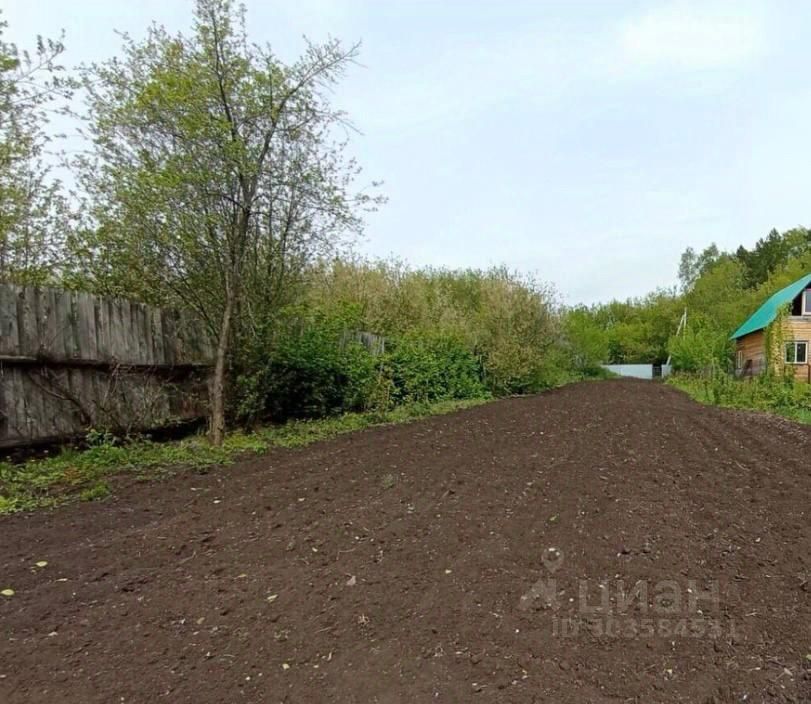 На фотографии изображен участок земли, подготовленный для посадки. Земля рыхлая и ровная, что создает благоприятные условия для выращивания растений. С одной стороны участка расположена деревянная заборная конструкция, а с другой виднеется деревянный дом с зеленой крышей. Вокруг участка растут деревья и кустарники, создавая уединенную атмосферу. Участок окружен зелеными насаждениями, что придает месту природный вид. Общая композиция участка свидетельствует о возможности для садоводства и ландшафтного дизайна.