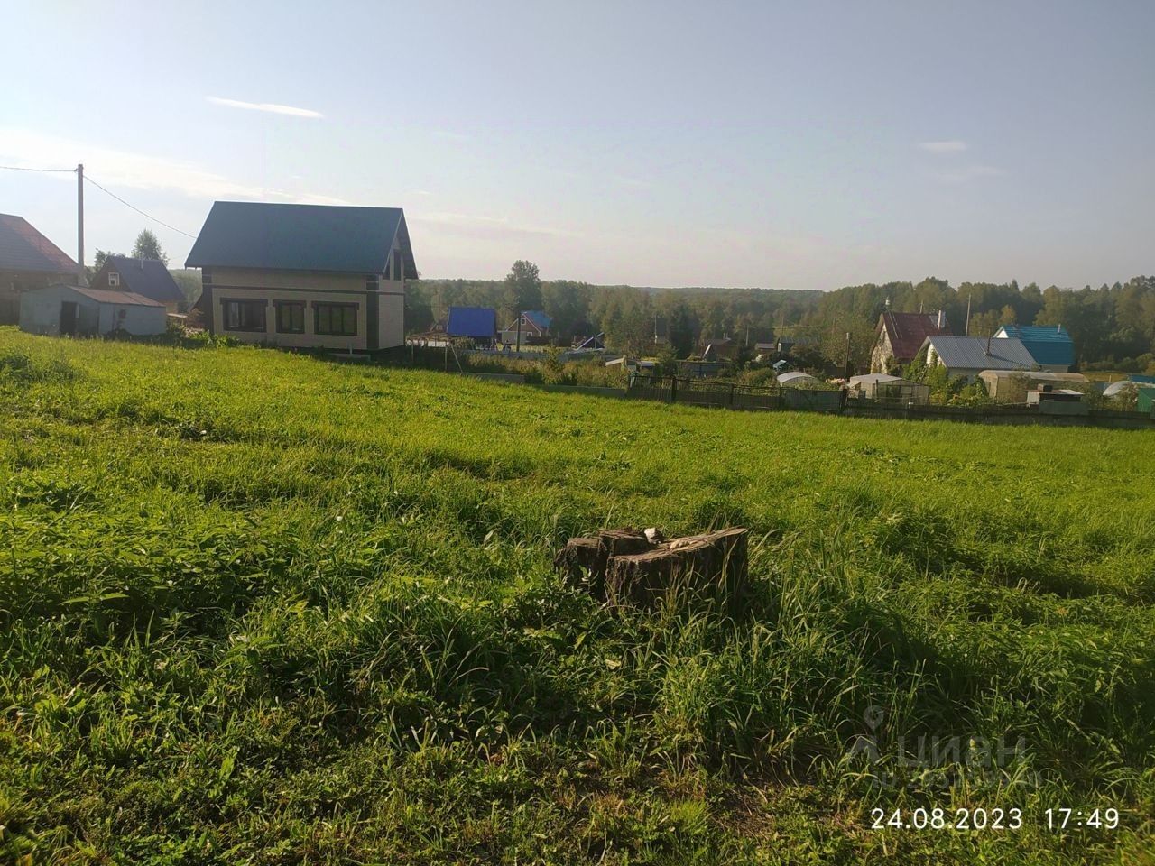 На фотографии представлено загородное строение в современном архитектурном стиле с наклонной крышей и большими окнами, обеспечивающими естественное освещение. Стены выполнены из светлого материала, что создает ощущение простора. Вокруг дома ухоженный зеленый газон с высокой травой, что придает участку живописный вид. Видны соседние постройки, выполненные в различных цветах, что создает разнообразие в ландшафте. Основные цвета – светлые и натуральные, что гармонирует с окружающей природой.