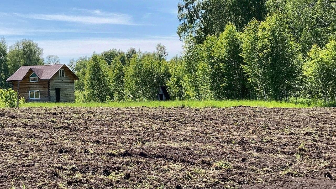 На фотографии представлено деревянное загородное строение в деревенском стиле с двускатной крышей, покрытой черепицей. Стены выполнены из бревен, что придает дому естественный вид. Окна имеют небольшие размеры, что характерно для традиционной архитектуры. Вокруг дома виден ухоженный участок с ровной почвой, готовой для посадки, а также зеленые деревья на заднем плане, создающие уютную атмосферу. Цвета в основном теплые и естественные, с преобладанием коричневых и зеленых оттенков, что подчеркивает гармонию с природой.