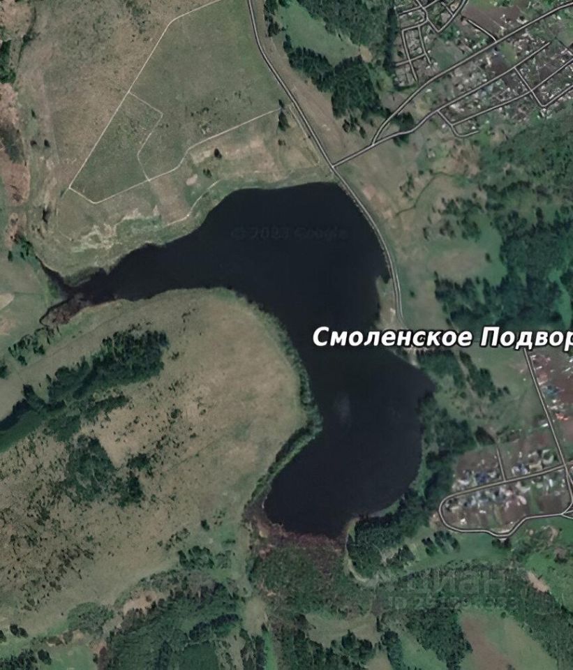 Вид на водоем с окружающими его зелеными ландшафтами. На фотографии заметны участки земли, обрамляющие озеро, с различной растительностью. Вдоль береговой линии видны небольшие лесные массивы и открытые поля. Параллельно водоему проходит дорога. Структура местности имеет холмистый рельеф, создавая живописные виды. Вдали можно разглядеть застройку, но детали зданий не различимы. Цветовая палитра включает насыщенные зеленые и коричневые оттенки, подчеркивающие природную красоту этого района.