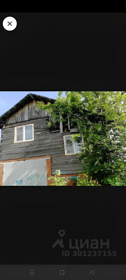 Здание выполнено в традиционном деревянном архитектурном стиле с использованием тёмной обшивки. На фасаде видны окна с простыми рамами, а также элементы, свидетельствующие о старинной постройке. Структура имеет два этажа, что видно по расположению окон и общему дизайну. Основные материалы – дерево и кирпич, придающие конструкции rustic-атмосферу. Цветовая гамма включает тёмные оттенки дерева и светлые кирпичные элементы. Отсутствие отделки на некоторых участках указывает на необходимость ремонта. Растительность вокруг добавляет естественности и уюта к общему виду здания.