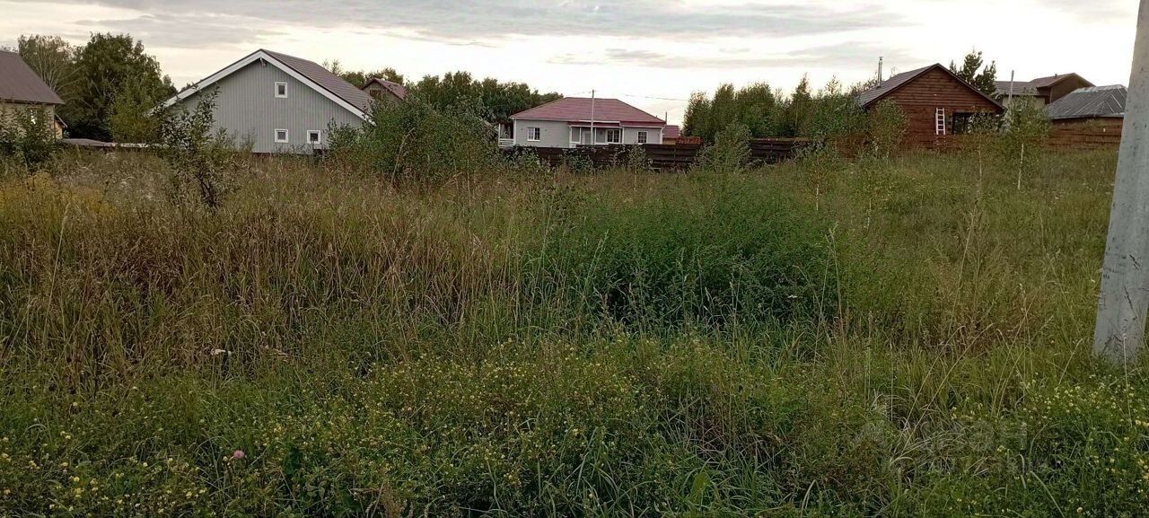 На фотографии виден участок земли с высокой травой и редкими кустарниками. На заднем плане расположены несколько домов с разной архитектурой: один из них с серыми стенами и двускатной крышей, другой выполнен из дерева с традиционным дизайном. Участок выглядит неухоженным, с дикой растительностью, что создает ощущение естественной среды. Цвета земли и растительности варьируются от зеленого до коричневого, а строения имеют нейтральные оттенки, что гармонирует с окружающей природой.