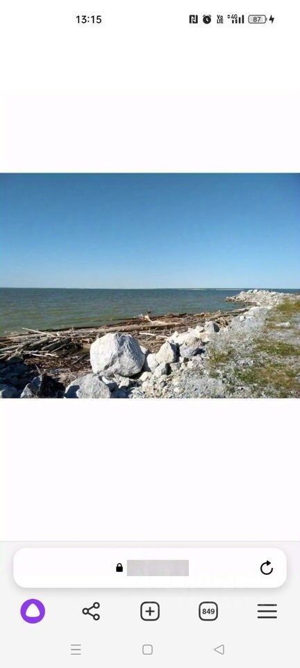 На изображении виден берег водоема с валунами и древесными остатками. Вода спокойная, с легкими волнами, отражающими ясное небо. Песчаная и каменистая поверхность прибрежной зоны создает естественный переход от воды к суше. Видны большие камни, разбросанные по берегу, а также древесные бревна, образующие неформальные линии. Общая атмосфера спокойная и умиротворяющая, подчеркивающая природную красоту данного места.