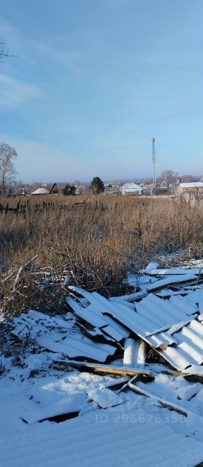 На фото изображен участок земли, покрытый снегом. Видны остатки строительных материалов, таких как металлические листы и древесина, разбросанные по земле. Участок окружен сухими кустарниками и травой, создающими ощущение заброшенности. На заднем плане виднеются несколько домов, построенных в традиционном стиле, с крышами, покрытыми снегом. Присутствуют элементы сельской архитектуры, такие как заборы и деревья, что подчеркивает атмосферу загородной местности. Цветовая палитра состоит из холодных оттенков белого и серого, создающих зимнее настроение.