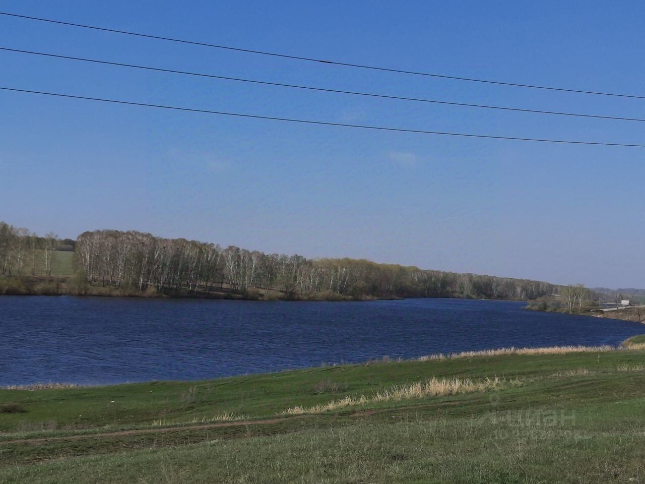 На фотографии видно спокойное водное пространство с широким берегом, покрытым зелёной травой. Вдали расположены деревья, создающие естественную границу между водой и открытым пространством. Вода выглядит чистой и спокойной, отражая свет голубого неба. Линия горизонта плавно изгибается, добавляя глубину изображению. Цвета преобладают натуральные: зелёный, синий и коричневый, что создаёт гармоничное и умиротворяющее впечатление. Пространство выглядит идеальным для отдыха на природе или водных активностей.