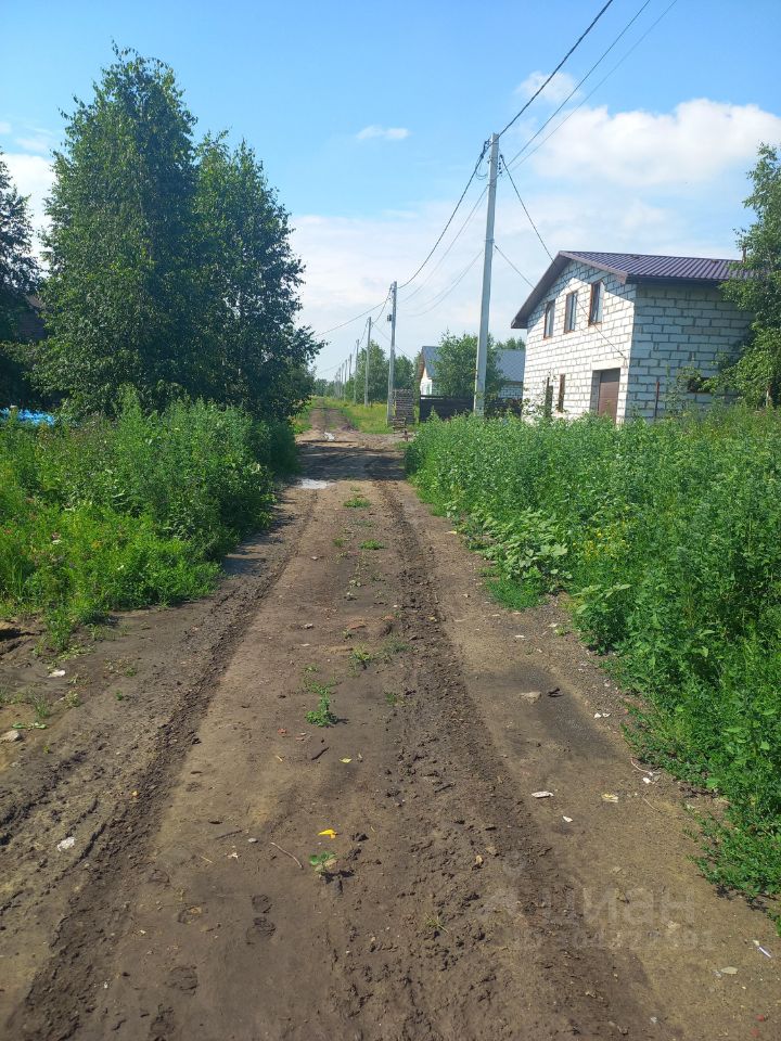 На фотографии виден грунтовый путь, окруженный высокой зеленью и деревьями. Дорога имеет неравномерную поверхность с следами от колес. С одной стороны дороги расположены дома, построенные из светлого камня с темными крышами. Структурные элементы включают окна и двери, которые придают зданиям простой, но функциональный вид. Вдоль дороги видны электрические столбы, что указывает на наличие коммуникаций. Общая атмосфера создает ощущение уединенности и близости к природе.