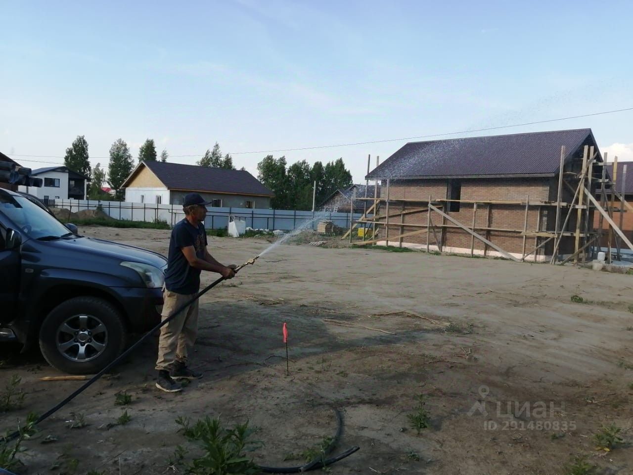 На фотографии видна строительная площадка с рабочим, который поливает землю. Стены зданий выполнены из кирпича, крыши имеют двускатную форму с темной черепицей. Вдали расположены несколько строений, одно из которых уже завершено, а другое находится на стадии строительства с деревянными опорами. Земля вокруг выглядит неухоженной, с небольшими растениями и участками голой почвы. Основные цвета – коричневый кирпич и темно-серая крыша. Рабочий одет в удобную одежду и использует шланг для полива.