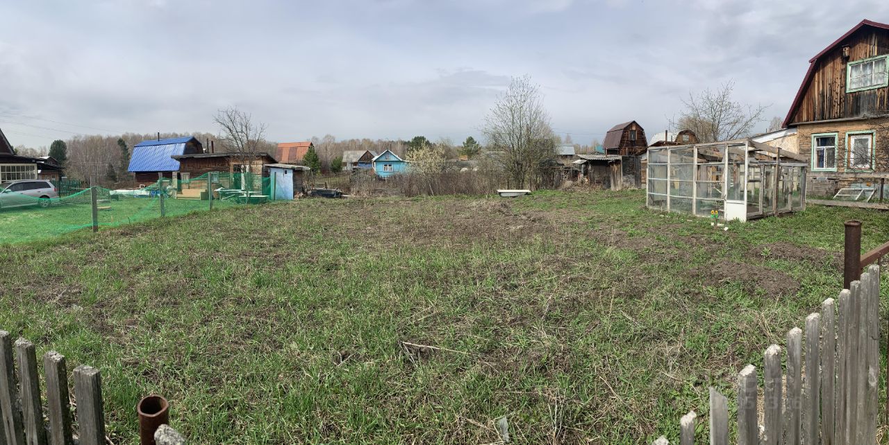 На фотографии виден просторный участок земли с ухоженной травой. В центре расположено пустое пространство, готовое для посадки. Слева находятся деревянные постройки с крышей из синего металла и характерными для дачных участков элементами. Справа виден стеклянный тепличный комплекс, который может использоваться для выращивания растений. Забор из деревянных досок ограничивает участок, добавляя уюта. Общая атмосфера участка спокойная и располагающая к отдыху на природе.