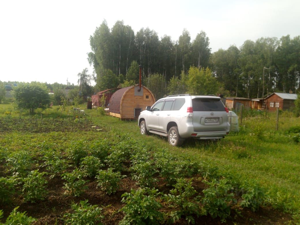 На фотографии представлено загородное строение, выполненное в виде деревянного бани с округлой формой и крышей, покрытой металлочерепицей. Внутри видна уютная атмосфера, характерная для русских бань. Окружение состоит из зелёного сада с грядками, где растут картофель и другие растения. На переднем плане расположена серебристая машина, что указывает на доступность и удобство для владельцев. Деревянные постройки на заднем плане создают гармоничное сочетание с природой. Основные цвета – зелёный, коричневый и серый, что придаёт месту естественность и спокойствие.