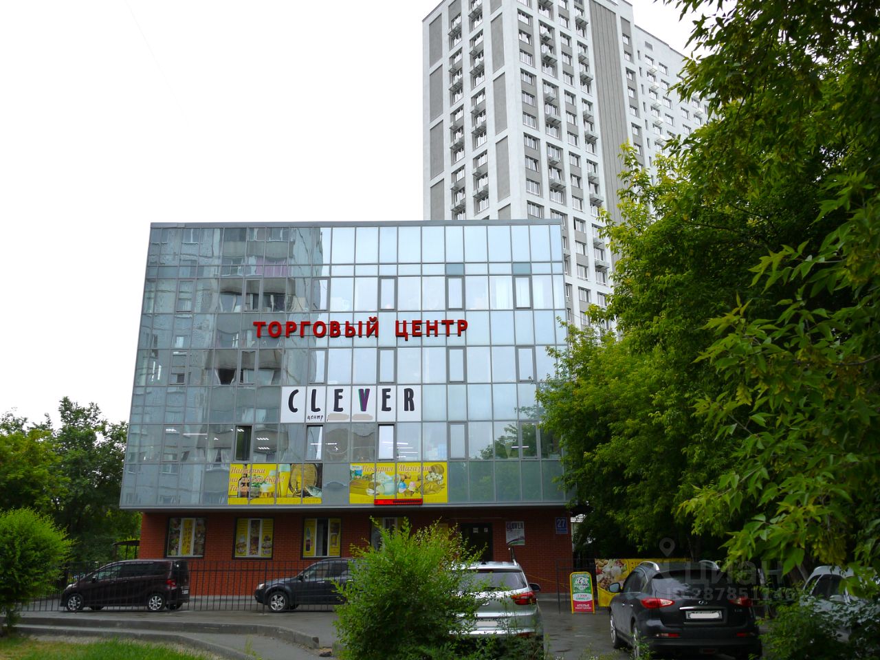 Современный торговый центр с стеклянным фасадом, отражающим окружающие здания. Архитектурный стиль минималистичный, с акцентом на прозрачность и свет. На первом этаже расположены витрины и входные двери, оформленные в ярких цветах. Главный элемент - крупные красные буквы, обозначающие название заведения. Стены выполнены из стекла и кирпича, создавая интересный контраст. Цветовая палитра включает нейтральные тона, а также яркие акценты на рекламных вывесках. Общее впечатление - современное и привлекательное пространство для покупателей.