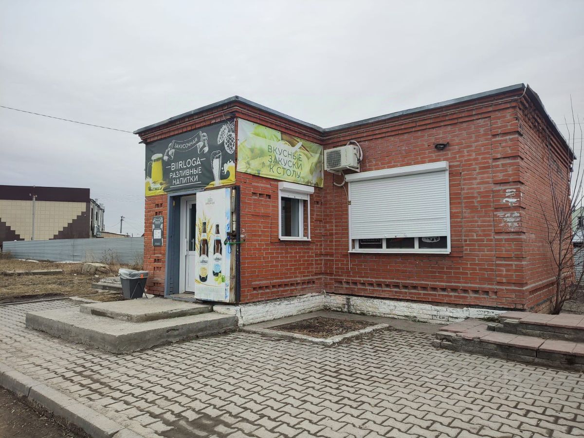 Здание выполнено в кирпичном архитектурном стиле с элементами современного дизайна. Структура включает в себя небольшую одноэтажную постройку с прямоугольной формой и плоской крышей. На фасаде видны окна с защитными рольставнями и вывеска с яркими графическими элементами. Вход оформлен ступеньками, ведущими к двери. Основные материалы – красный кирпич и бетон. Цветовая гамма включает в себя натуральные оттенки кирпича и яркие акценты на вывеске. Финишные детали, такие как кондиционер и освещение, добавляют функциональности и современности.